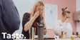 A woman with blonde hair tastes a dark beverage from a black travel mug, with another woman drinking from a glass at the table beside her. The word 'Taste.