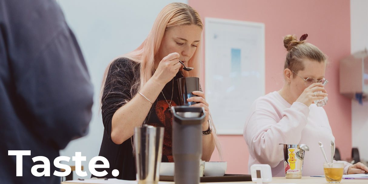 A woman with blonde hair tastes a dark beverage from a black travel mug, with another woman drinking from a glass at the table beside her. The word 'Taste.