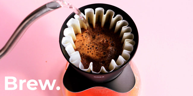 A close-up of a pour-over coffee setup with hot water being poured from a metal gooseneck kettle into a black dripper filled with coffee grounds and white paper filter. The word 'Brew.
