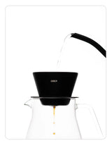 Coffee brewing setup with a OREA 01 Obsidian coated steel brewer and clear glass carafe on a white background