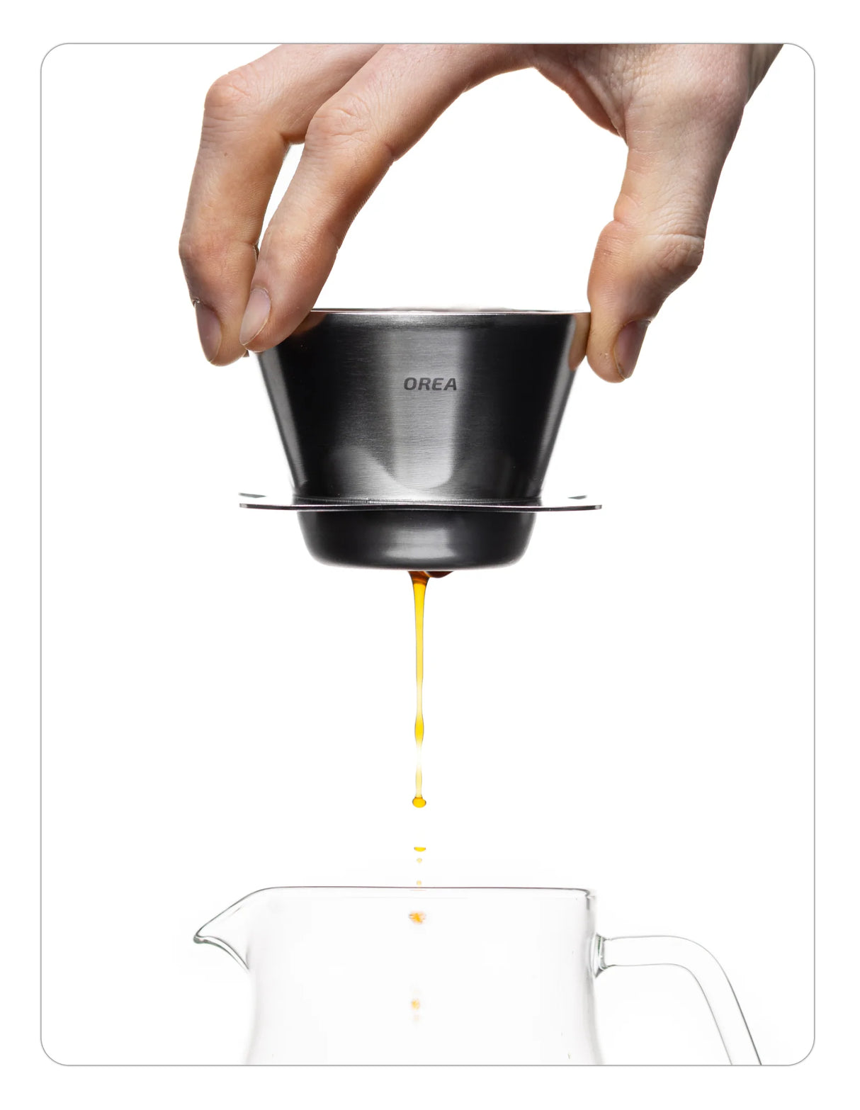 Hand pouring coffee from an OREA 01 raw steel coffee dripper into a glass pot on a white background